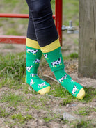 Vibrant unisex cow socks with black and white cows wearing yellow wellies on a grass green background and bright yellow top, heel and toe. 